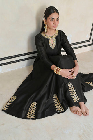 Model in a black traditional outfit with gold embroidery sitting on floor.