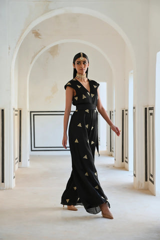 A woman models a black floor-length jumpsuit with gold embroidered motifs in a white arched corridor.