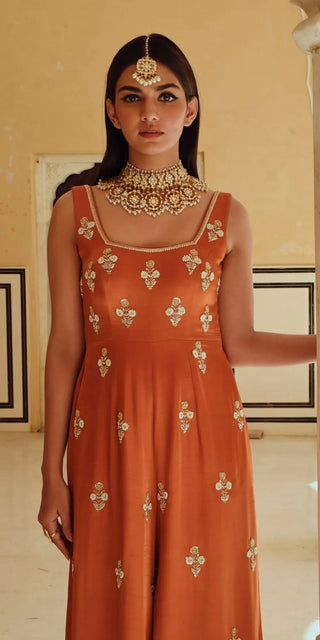 A woman wearing a sleeveless rust-orange Jumpsuit with gold floral embroidery, styled with traditional Indian jewelry, standing in an indoor setting with beige walls.