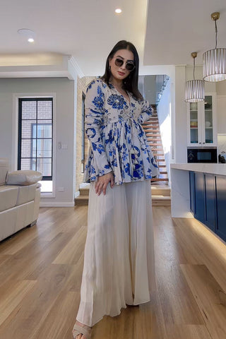 Woman in a blue floral peplum top and ivory palazzo standing in a modern living room. This ethnic chic peplum dress is designed by Paulmi and Harsh.