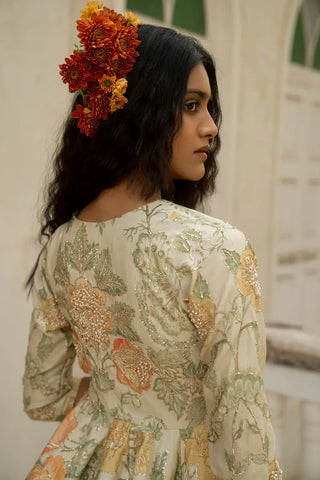 A woman shown from the back wearing a ivory-colored embroidered dress with green and orange floral patterns and a matching orange flower headpiece, set against a soft-focus white architectural background.