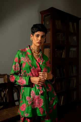 A model wearing a vibrant pnh floral green print silk blazer style co-ord set., standing in a dimly lit room with a dark wooden bookshelf in the background.