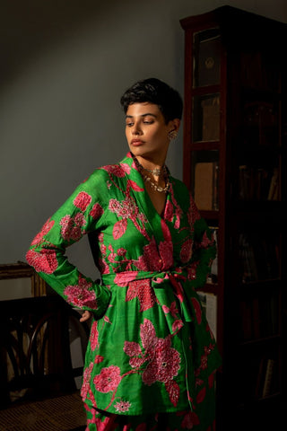 A woman wearing a vibrant green kimono-style robe with a bold pink floral pattern, standing in a dimly lit room with a dark wooden bookshelf in the background.