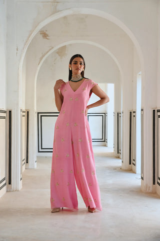A woman stands in a white arched corridor wearing a vibrant pink indo-western jumpsuit adorned with delicate gold butti embroidery — a modern silhouette that celebrates the beauty of traditional craftsmanship. Perfect for the woman who loves contemporary fashion with an ethnic touch.