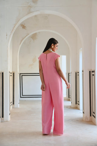 A woman stands in a white arched corridor wearing a vibrant pink jumpsuit posing the show the back of Jumpsuit.