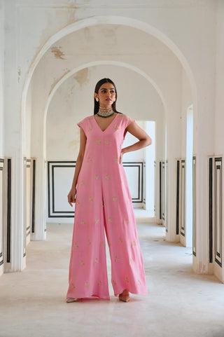 A woman stands in a white arched corridor wearing a vibrant pink wide-leg jumpsuit adorned with delicate gold butti embroidery. She poses to highlight its effortless elegance — perfect for occasion wear, wedding cocktail nights, and festive celebrations with family and friends.