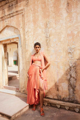 A model posing in a resort look. The dress is in coral colour made from linen crop top, Linen jacket with cut work floral embroidery, silk organza warp pre-stitched skirt. A perfect resort look book dress, holiday, vacation, coastal, beach wear.