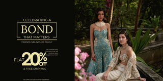 Promotional banner for a fashion brand featuring two models in traditional Indian attire. On the left, a model in a light blue embroidered jumpsuit stands, while on the right, another model in a cream and gold embroidered Jacket set on a stone ledge surrounded by greenery and pink flowers.