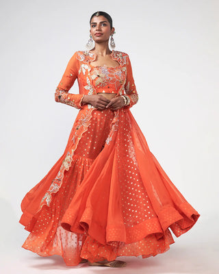 A woman modeling a bright orange traditional Indian dress crafted in organza and georgette with applique hand work and patch work detailing on cape, blouse. The . set against a plain light gray studio background. Narangi Dhadak Set is perfect dress style for wedding and bridal occasions.