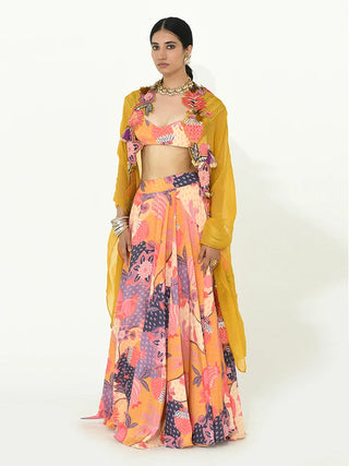 A model wearing a vibrant Indian ethnic wear set featuring a multicolor floral print lehenga choli with a sheer mustard yellow dupatta, set against a plain white studio background.